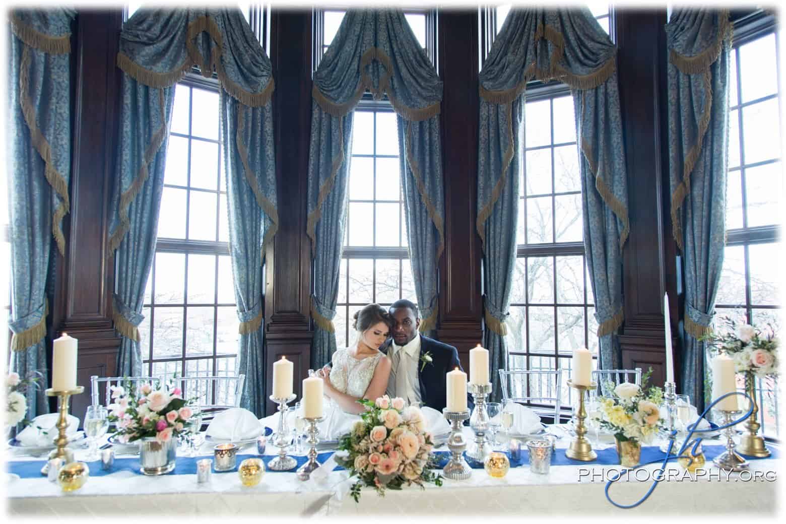 A bride and groom sit closely together at an elegantly decorated reception table with large windows and blue drapes in the background.